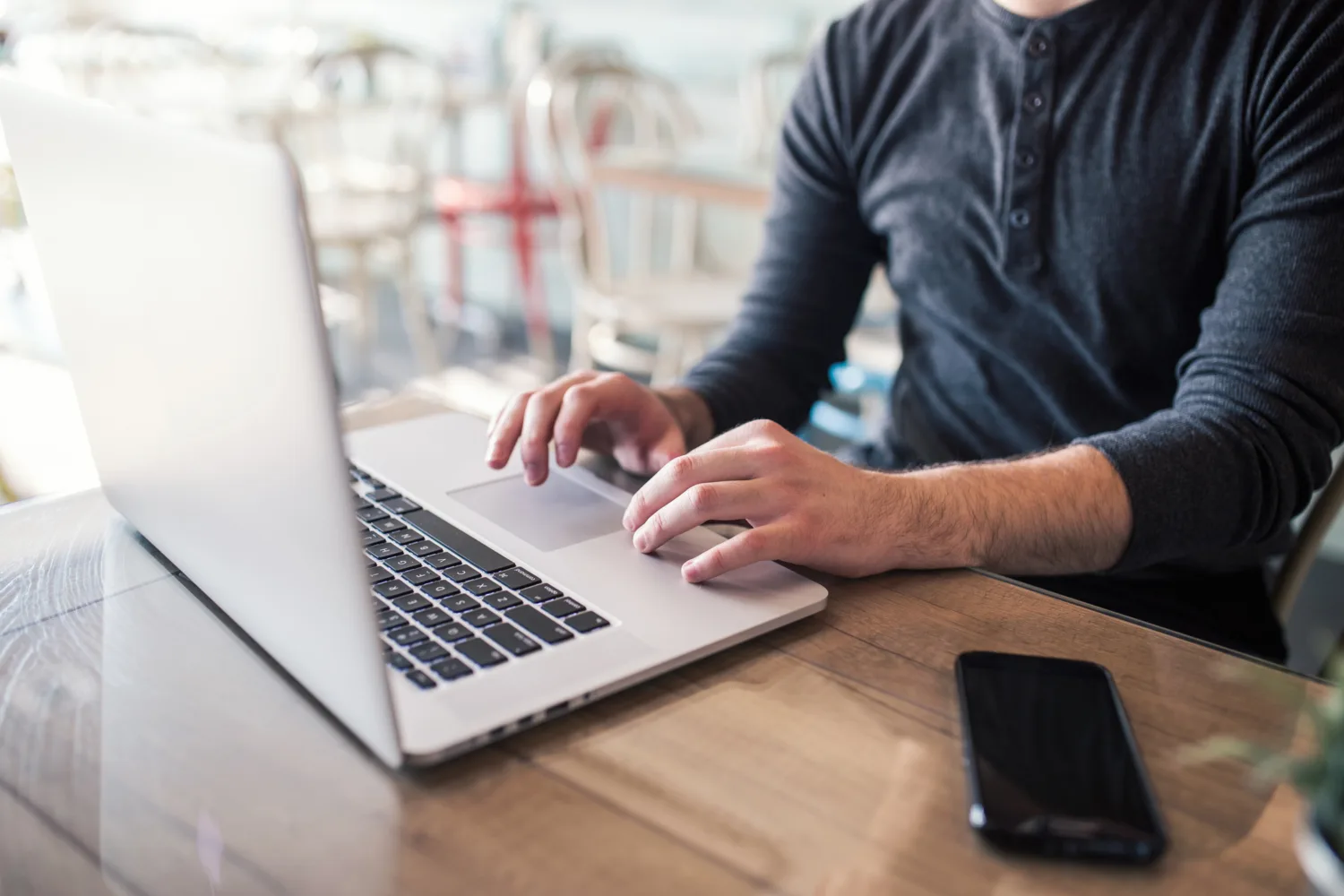 A man's hands are seen interacting with the touchpad on a laptop. He is sitting in a Cafe area.